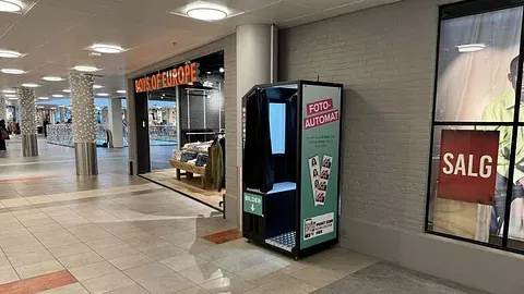 Fotoboks plassert i et kjøpesenter, med butikk i bakgrunnen.